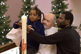 Laken Tchoutouo, 4, lights her sister&#039;s baptismal candle with the help of their father, Hypolyte Tchoutouo, and Deacon Simon Nadeau, during a Dec. 9 commemorative Mass at St.-Pierre-aux-Liens Church in Quebec City. Michelle Solaye Mane Tchoutouo, 8, was accidentally killed by a snowplow in Quebec City Dec. 1.