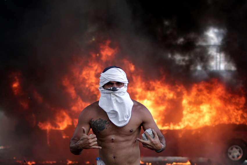 An opposition demonstrator gestures while carrying stones in front of a burning bus during a violent protest near Carlota airbase in Caracas, Venezuela, April 30, 2019. Armored vehicles plowed into anti-government protesters as troops loyal to Venezuelan President Nicolas Maduro tried to restore order; opposition leader Juan Guaido took to the streets in an attempt to lead a military uprising against the embattled president. 