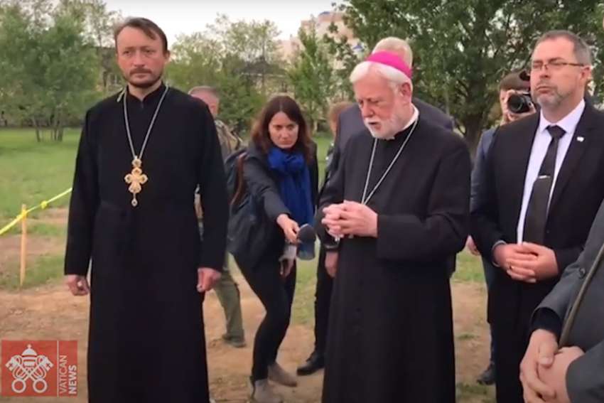 Archbishop Paul R. Gallagher, Vatican foreign minister, prays at the site of a mass grave near the Orthodox Church of St. Andrew in Bucha, Ukraine, May 20, 2022, in this still image taken from video posted by the Vatican. Pictured next to Archbishop Gallagher is Andrii Yurash, Ukraine&#039;s ambassador to the Holy See.