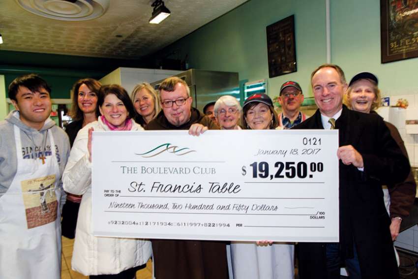 Br. John Frampton (centre, first row), animator of St. Francis Table, gets some help holding up a novelty cheque representing The Bouvlevard Club’s recent donation to the resturant for the poor. 