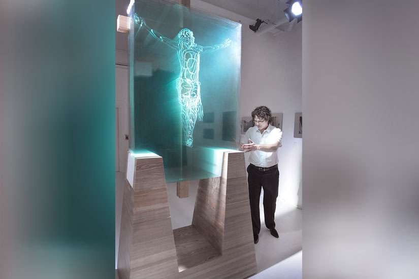 Jed Malitz poses for a photo with his work V2. The glass sculpture depicts the crucifixion and it is produced by light shining through 13 arranged panes of sculpted glass cut using a computer-guided process.