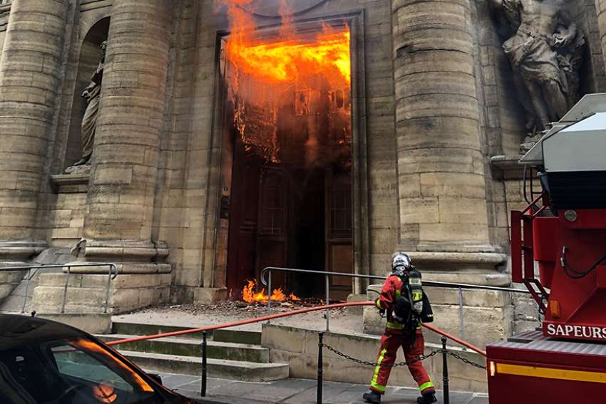 A firefighter is seen as flames shoot through the front door of St. Sulpice Church in Paris March 17, 2019, in this still image taken from social media. Vandals and arsonists have targeted French churches in a wave of attacks that has lasted nearly two months.