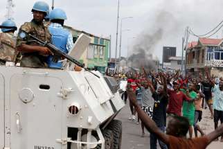 People chant slogans against Congolese President Joseph Kabila as armed U.N peacekeepers watch protesters Dec. 20, 2016 in Kinshasa.  In the Democratic Republic of the Congo, political unrest first erupted in 2015 after a bill was proposed which would potentially delay the presidential and parliamentary elections. The bill was widely seen by the opposition as a power grab on the part of Kabila.